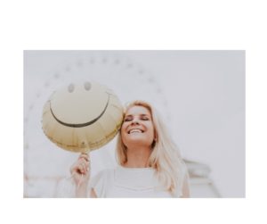 Joyful Lift: A woman smiling brightly as she holds a balloon adorned with a happy face, symbolizing light-heartedness and the uplifting power of a simple smile.