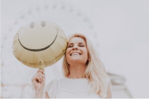 Joyful Lift: A woman smiling brightly as she holds a balloon adorned with a happy face, symbolizing light-heartedness and the uplifting power of a simple smile. 44 Simple Health Hacks.