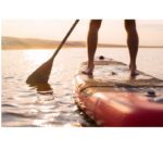 Serene Paddle Boarding: A person balanced on a paddle board glides across a calm lake, surrounded by the tranquil beauty of nature, illustrating an active yet peaceful outdoor pursuit for fitness and relaxation.