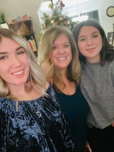 Image of Natalie, a middle-aged woman, smiling warmly alongside her two daughters in a cozy living room. Natalie is in the center, with one daughter on each side, all embracing each other. The background shows a tastefully decorated room, conveying a sense of warmth and family closeness in a home setting.