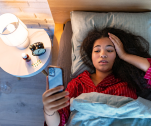 A tired woman lies in bed, looking at her phone, highlighting the common yet unhealthy habit that can interfere with adjusting to a time change.