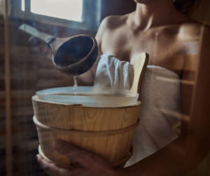 Woman in sauna pouring water from a wooden bucket onto hot stones to increase steam, wrapped in a towel and enjoying the relaxing heat.