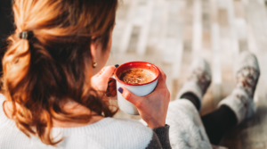 A Caucasian woman sitting outdoors, enjoying a hot beverage from a red mug, wearing a cozy knit sweater and socks. The focus is on the mug, capturing a moment of relaxation on a sunny day, with a blurred background adding a serene quality to the scene.