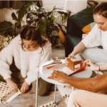 A diverse group of three people, including a Caucasian woman, a Caucasian woman with glasses, and a Black man, sitting around a small table in a festive living room, wrapping Christmas gifts together. The room is decorated with holiday elements like a small Christmas tree and potted plants, creating a cozy, festive atmosphere.