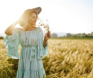 A woman in a flowing light blue dress and a wide-brimmed hat stands in a golden wheat field, gently holding wildflowers. The warm sunlight illuminates her peaceful expression, conveying a sense of freedom and connection with nature. Master Your Mind