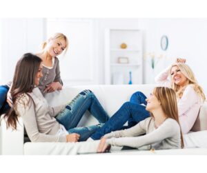 A group of four women relaxing and laughing together on a white sofa in a bright, modern living room, depicting strong social bonds and emotional intelligence.