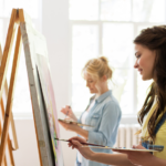 Women of various ages in a bright art studio, focused on painting on easels, expressing creativity in a community setting. Rediscover Your Passions.