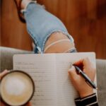 A woman sitting cross-legged on a couch, holding a coffee cup in one hand and writing in a journal with the other. She is wearing ripped jeans and a striped long-sleeve shirt, embodying a casual, reflective moment at home.