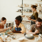 Four adults in a pottery workshop, collaboratively shaping and decorating ceramic wares, with shelves of pottery in the background.