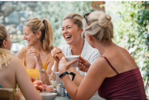 A group of women laughing and enjoying coffee together at an outdoor table, sharing a joyful moment in a relaxed setting. Rediscover Your Passions.