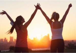 Two joyful women raising their arms in victory after a workout, with a sunset cityscape in the background, symbolizing triumph and personal wellness. Real Self-Help for Everyday Life!