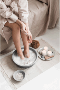 A woman in a soft pink robe soaking her feet in a basin with Epsom salts and bubbles, surrounded by candles and natural spa tools on a woven rug. Simple summer habits. 